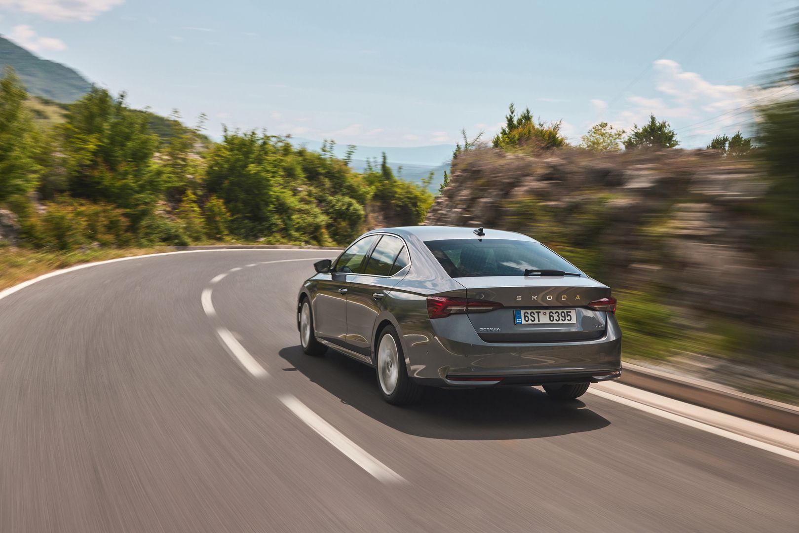 Silberner Skoda Octavia fährt auf kurviger Landstraße mit grüner Vegetation und Bergen im Hintergrund